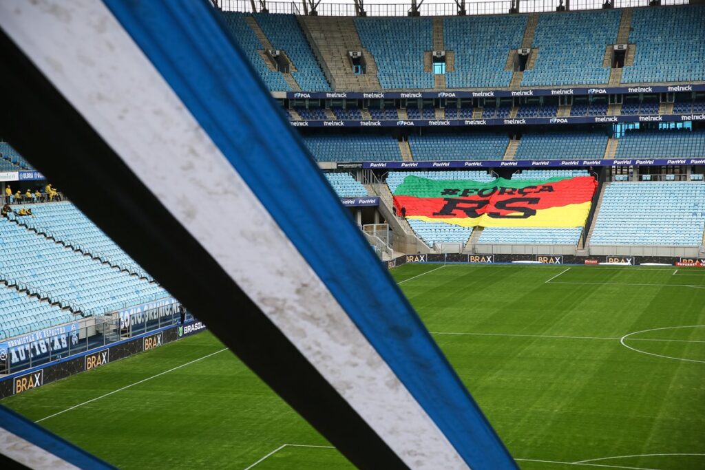 Imagem aberta da Arena do Grêmio; em primeiro plano uma faixa tricolor e na arquibancada ao fundo um mosaico com a bandeira do Rio Grande do Sul e os dizeres "#ForçaRS"